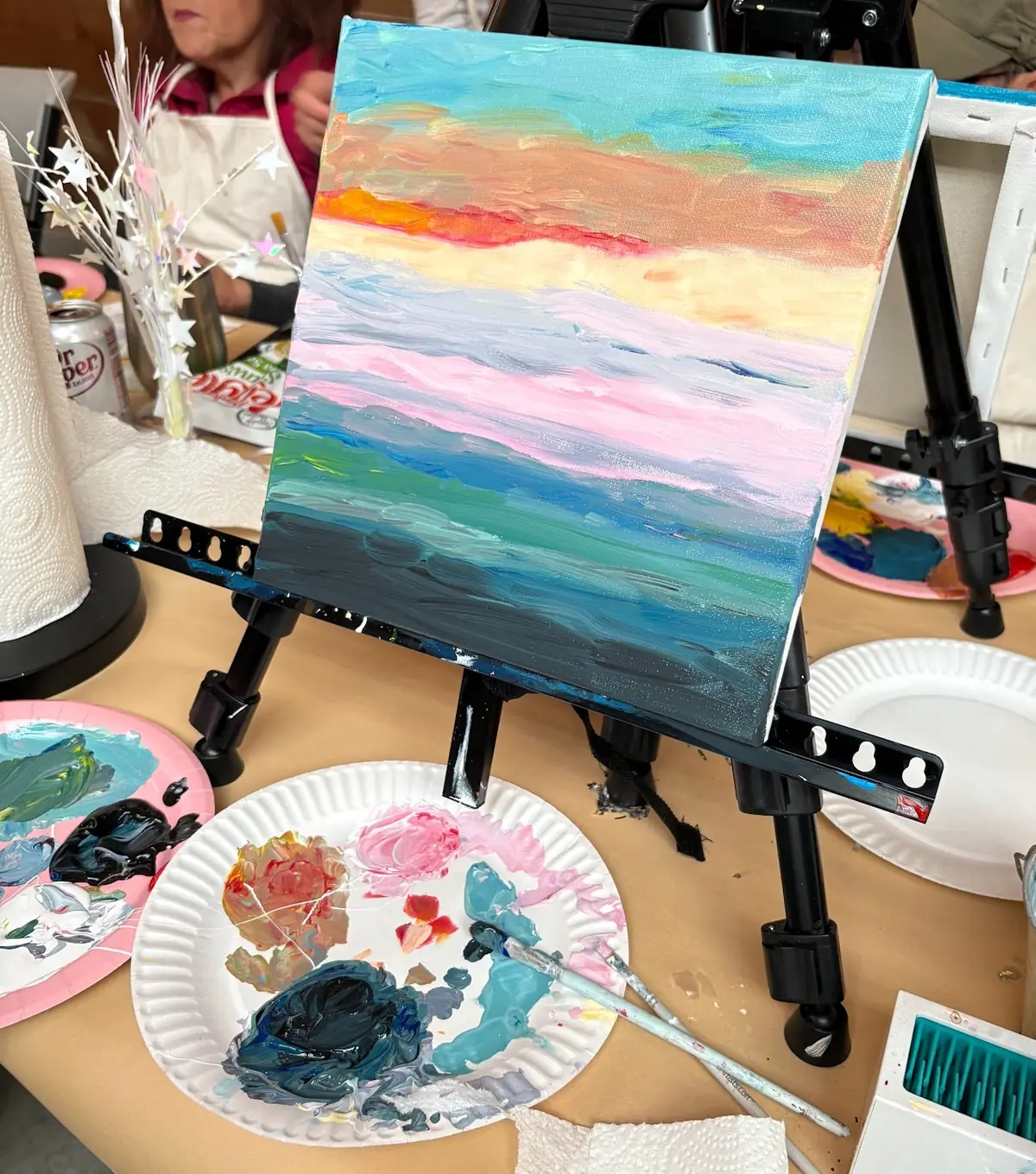 A colourful abstract sunset canvas beside a palette of paints