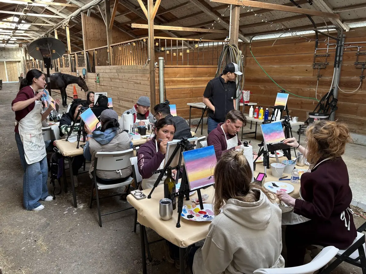 Friends at a painting table, the session's sample canvas on an easel