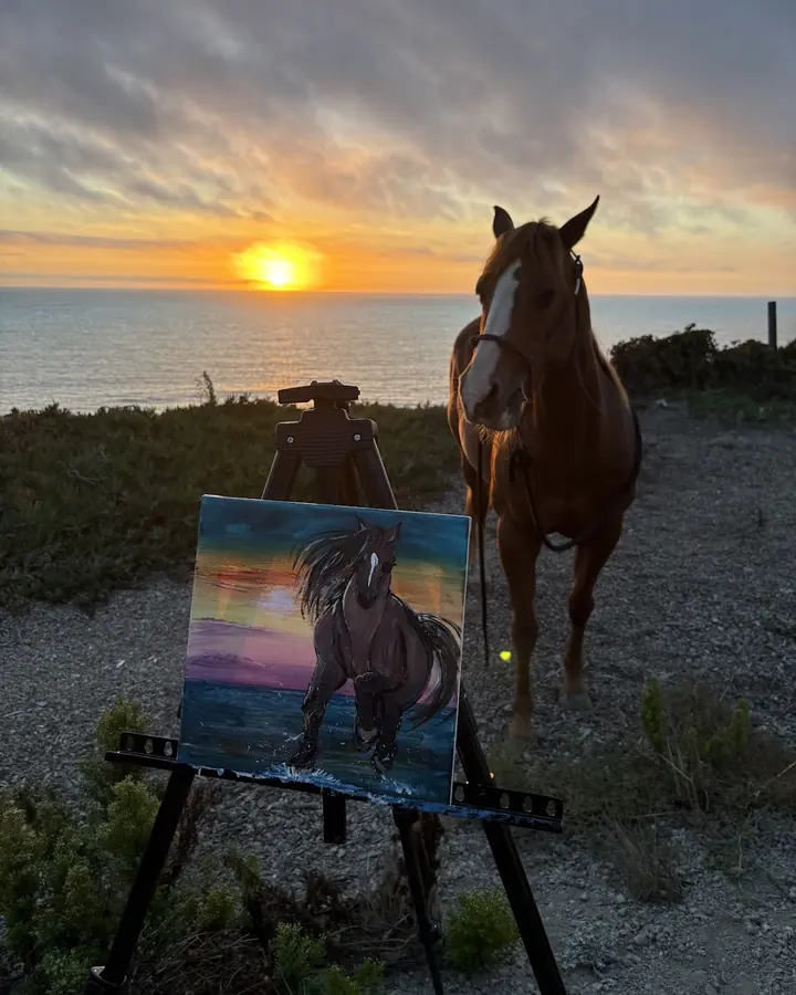A horse at sunset on the bluff beside an easel holding a painting of a running horse