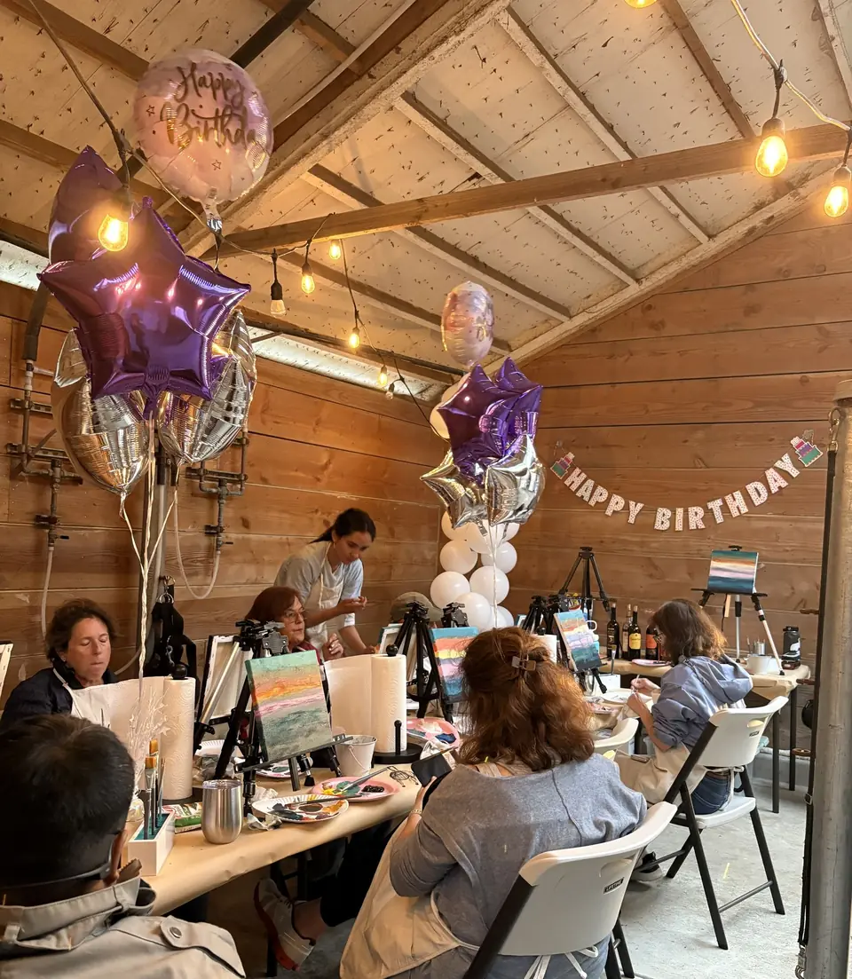 A birthday celebration at painting tables in the barn, balloons and a Happy Birthday banner overhead