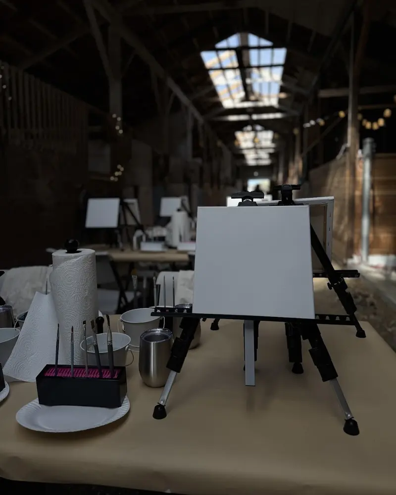 A blank canvas on an easel at a table set with paints, a paper towel roll and water cups